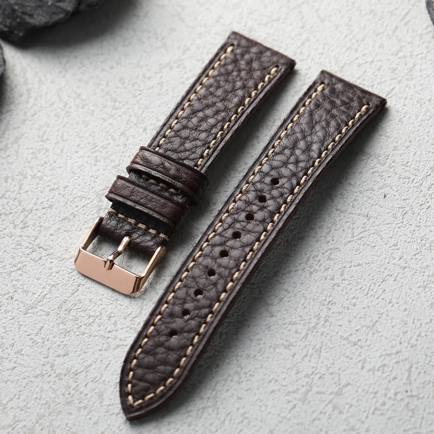 Brown leather watch strap with rose gold buckle on a light gray background