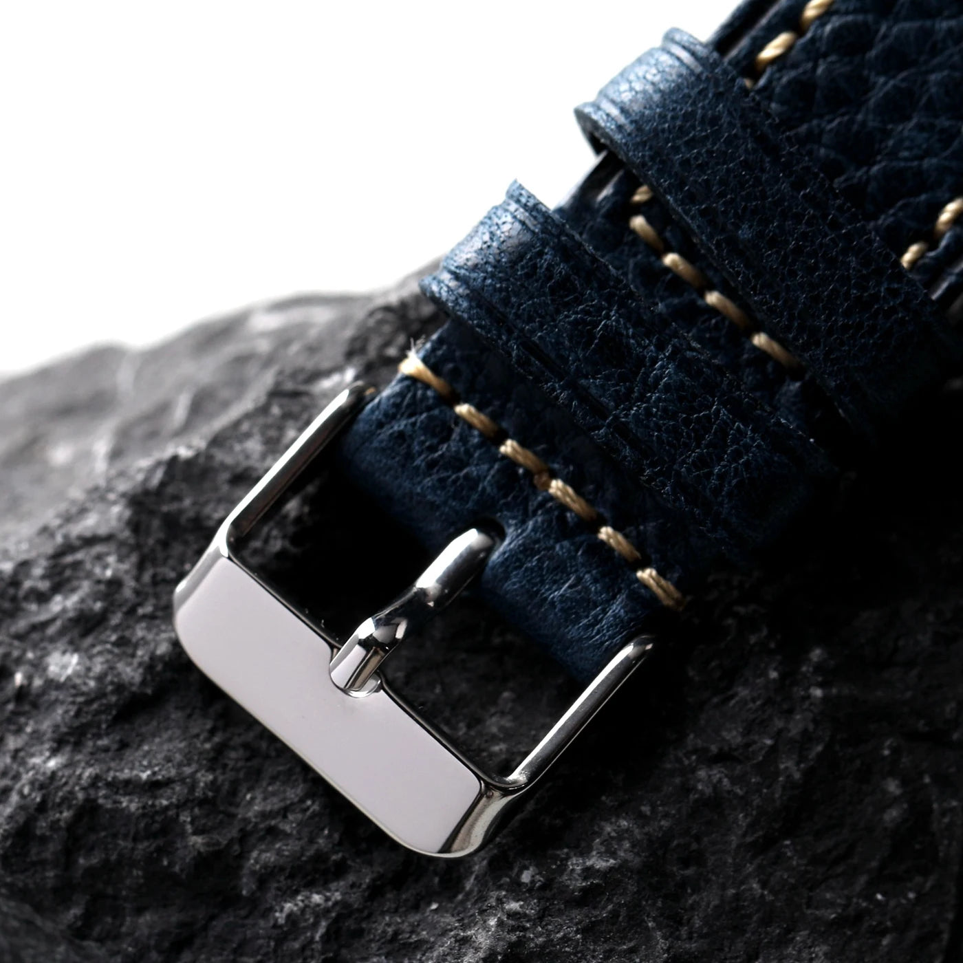 Close-up of a dark blue leather watch strap with a silver buckle on a rocky surface.