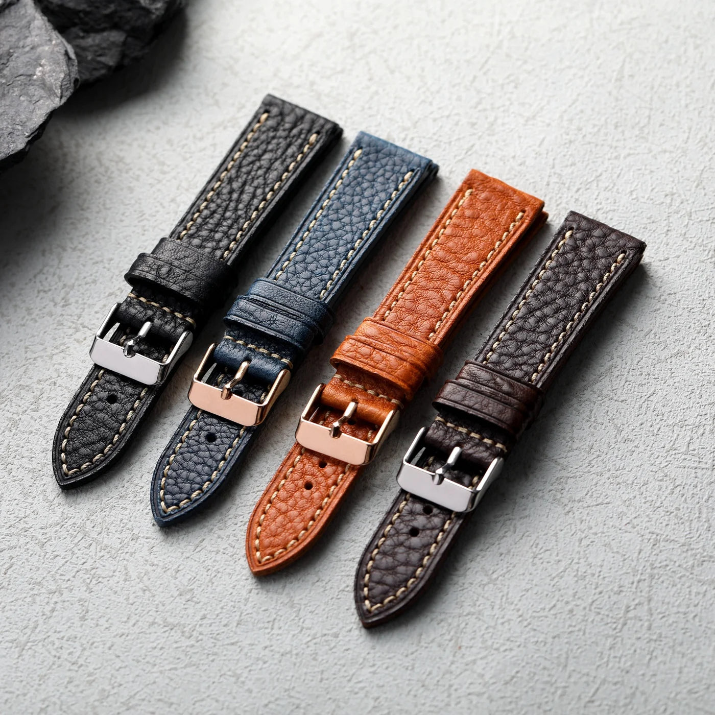 Four leather watch straps in black, blue, brown, and dark brown on a light gray surface.
