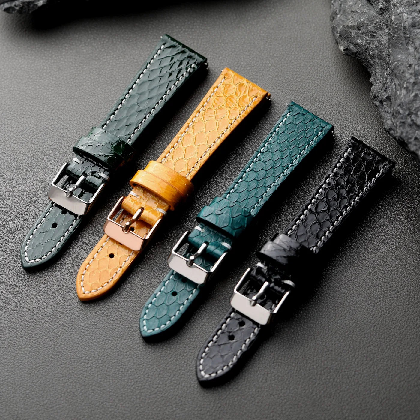 Python Snake Skin Four leather watch straps in black, green, yellow, and gray on a dark surface face up