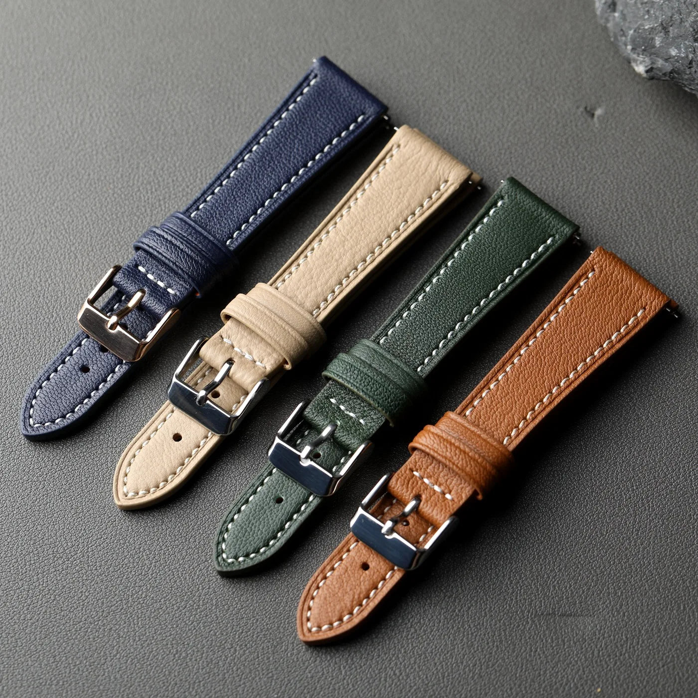 Four leather watch straps in blue, beige, green, and brown on a gray surface.