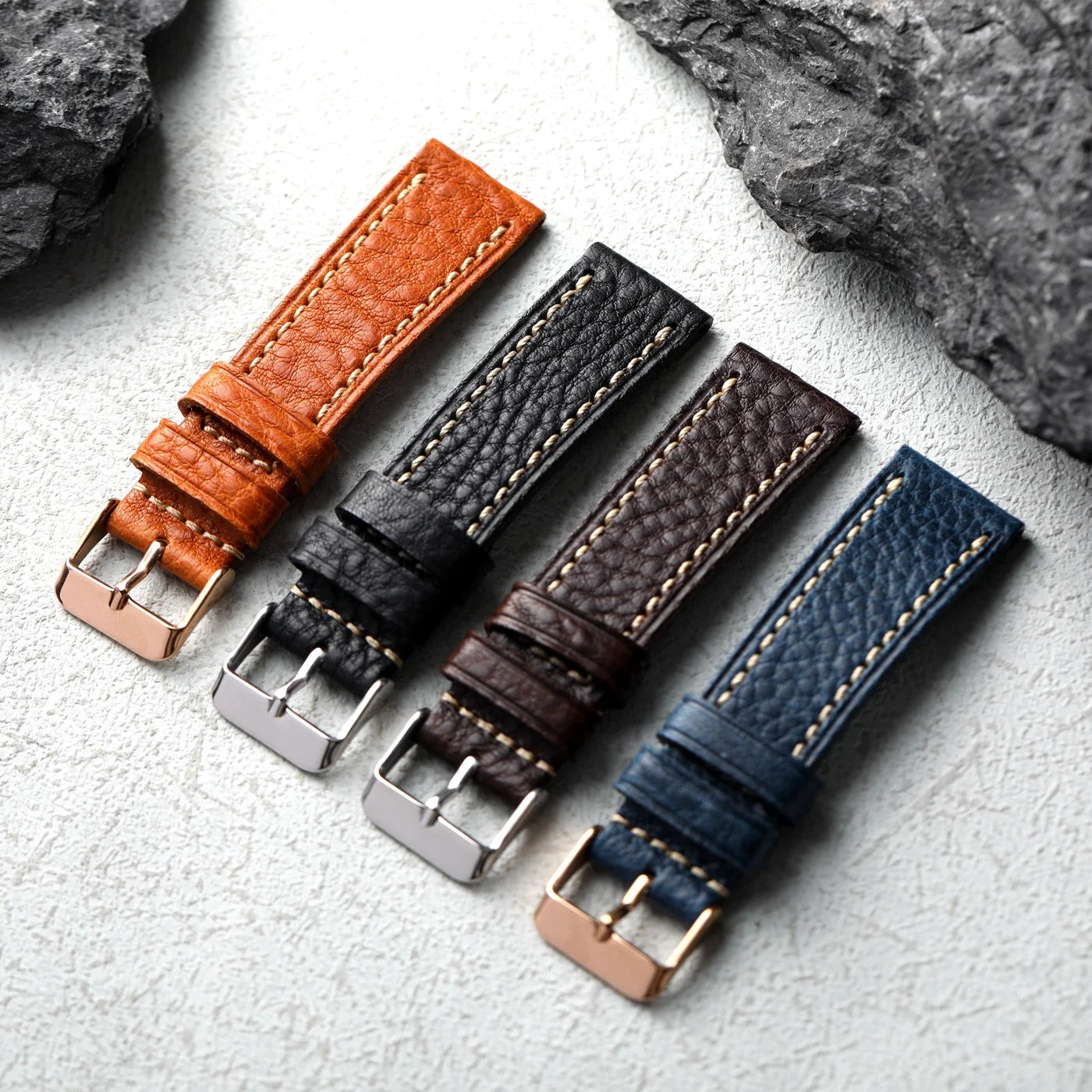 Four leather watch straps in brown, black, and blue on a textured surface.