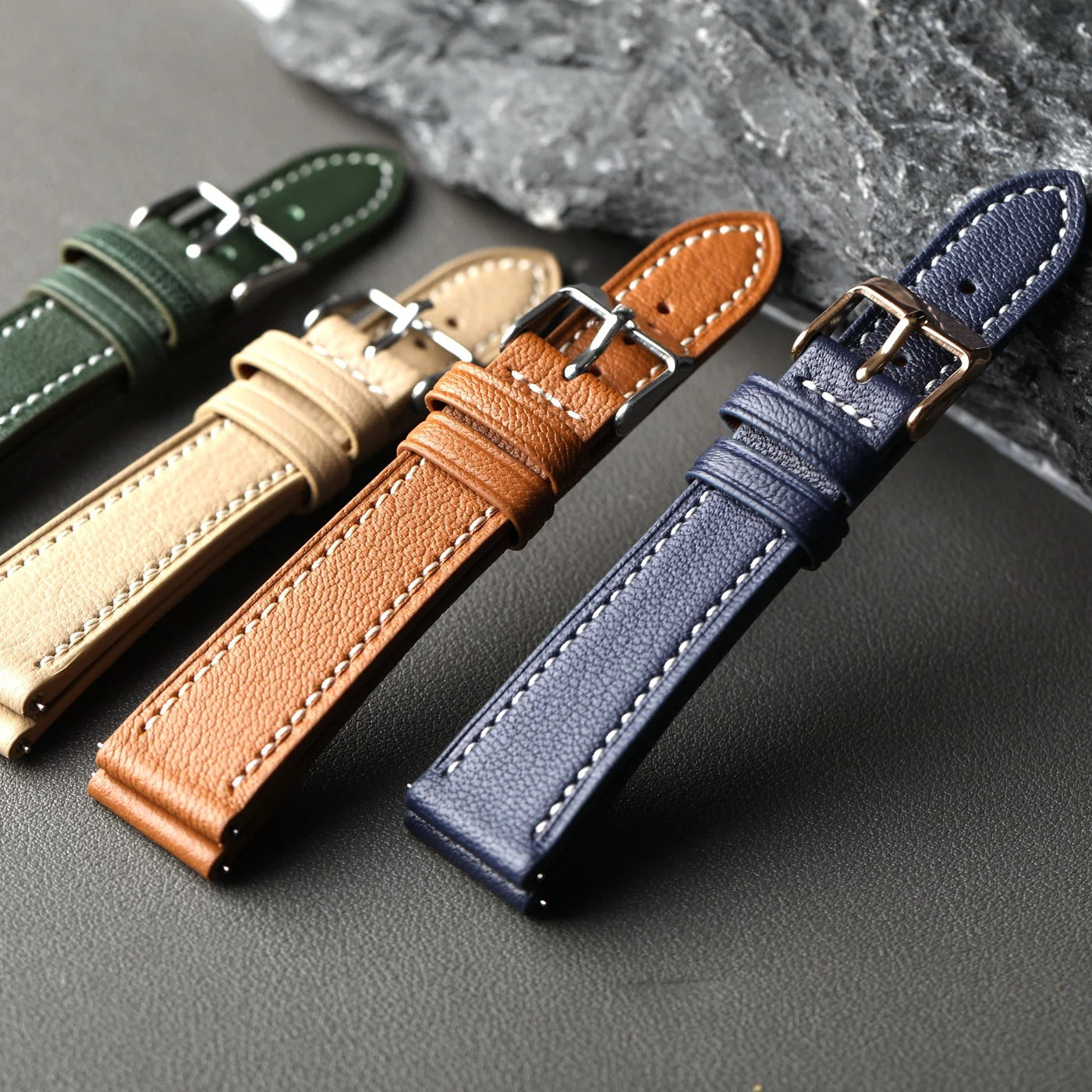 Four leather watch straps in green, beige, brown, and blue on a dark surface.