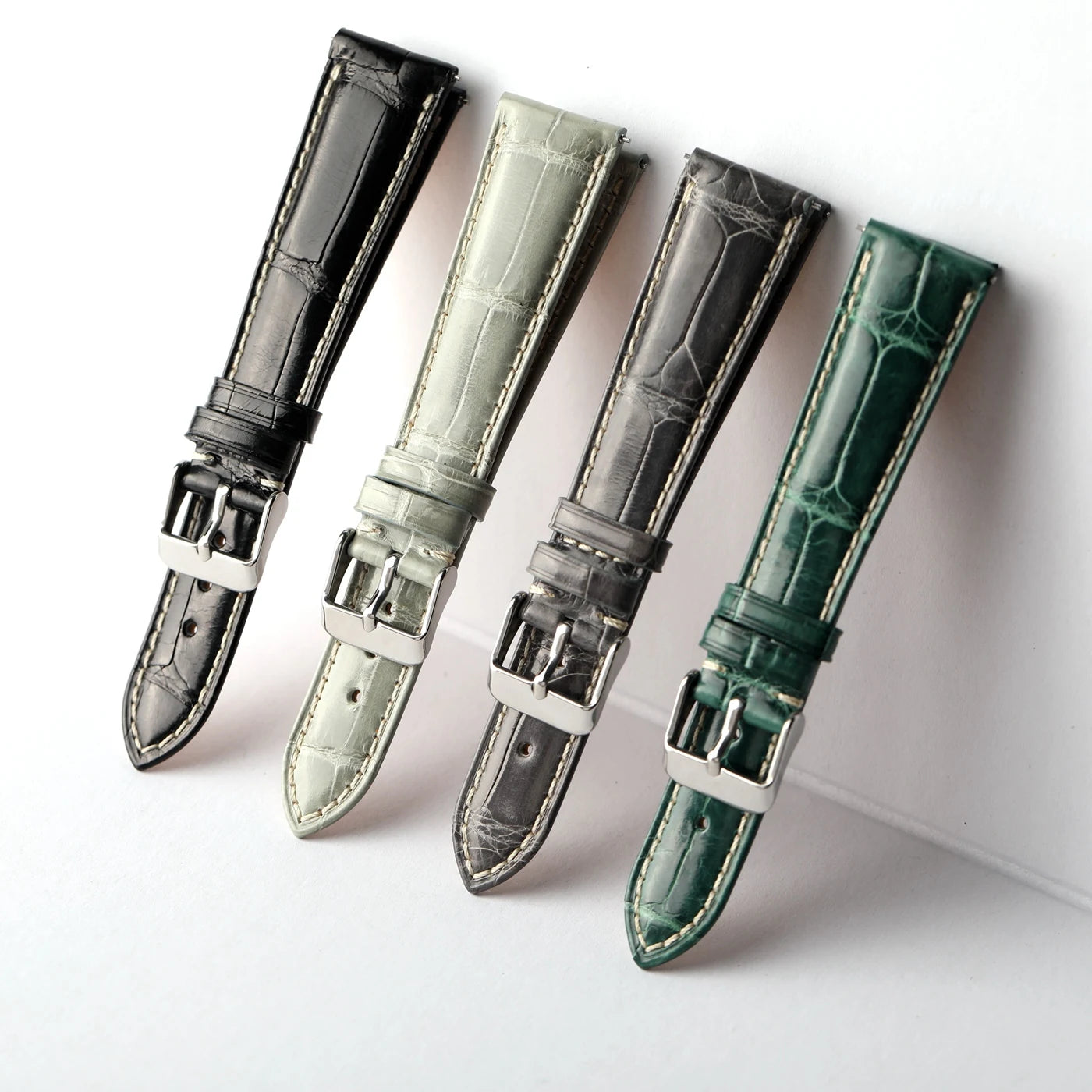 Four leather watch straps with silver-toned buckles arranged diagonally on a white background. The straps are black, light gray, charcoal, and forest green, each with contrasting stitching and an alligator or crocodile-embossed texture.