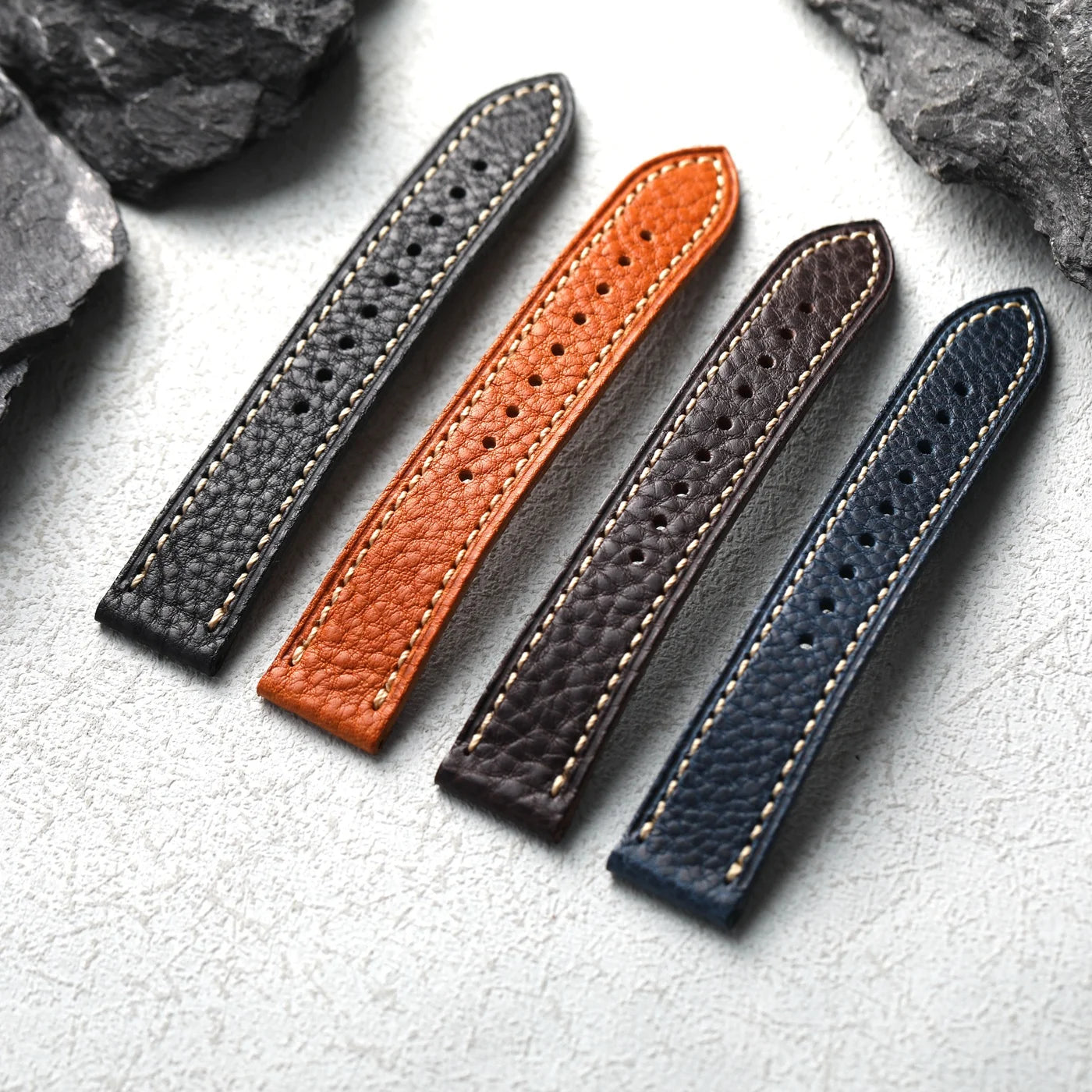 Four leather straps in gray, orange, brown, and blue on a textured surface with rocks in the background.
