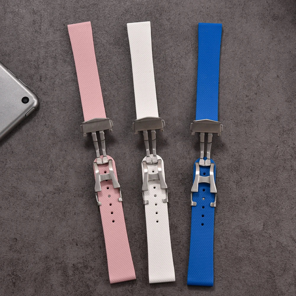 Three watch straps in pink, white, and blue on a gray surface with a phone partially visible.