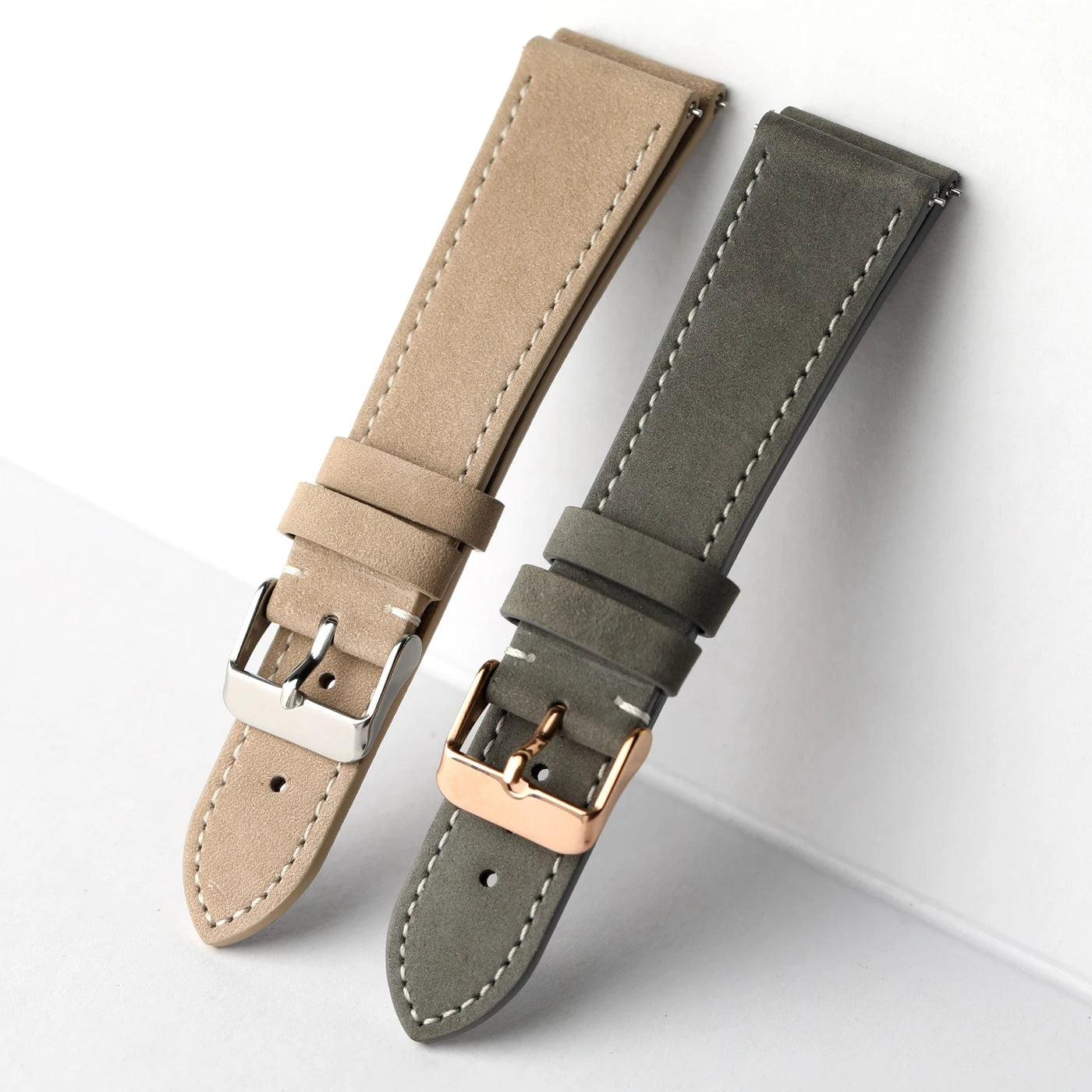 Two leather watch straps with white stitching, one in beige with a silver buckle and one in dark gray with a rose gold buckle, displayed on a light gray background.